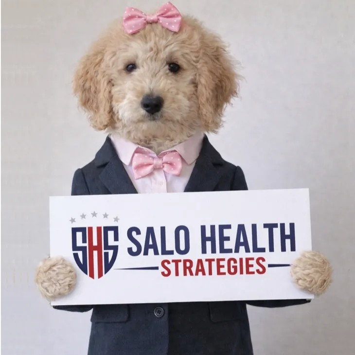 Misha Salo, a goldendoodle in a suit and pink bow tie, holding a Salo Health Strategies sign